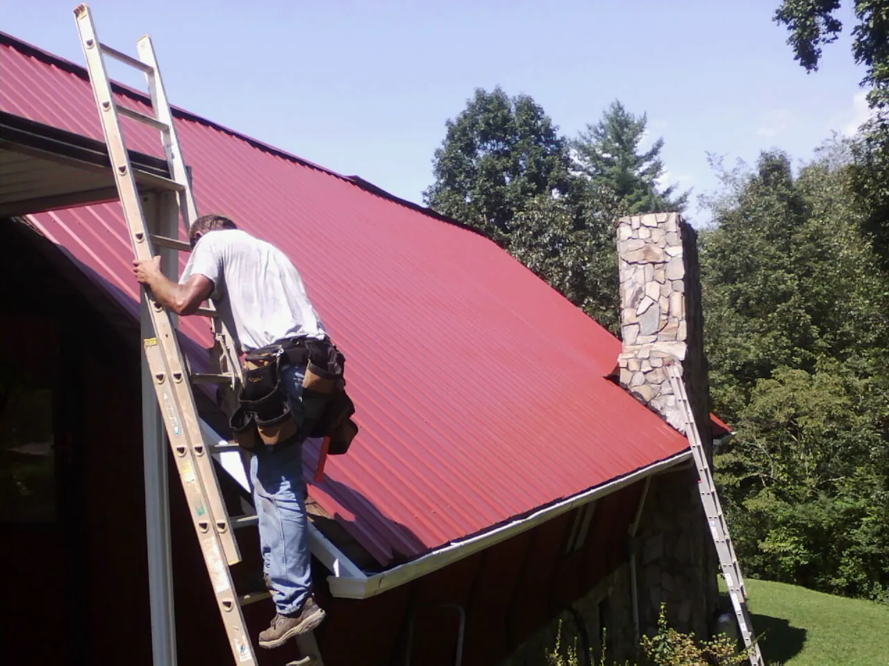 Roofer on a ladder beside a red metal roof and stone chimney — Soffit & Fascia Repair by Roofing Brothers in Park Hall